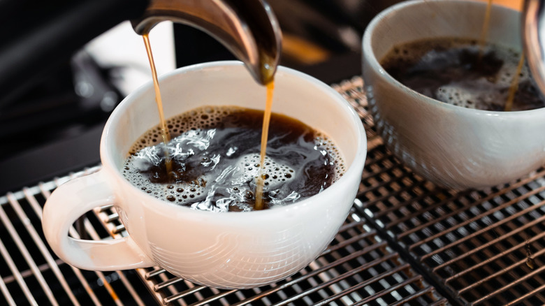 A white mug full of black coffee is being filled by a coffee machine