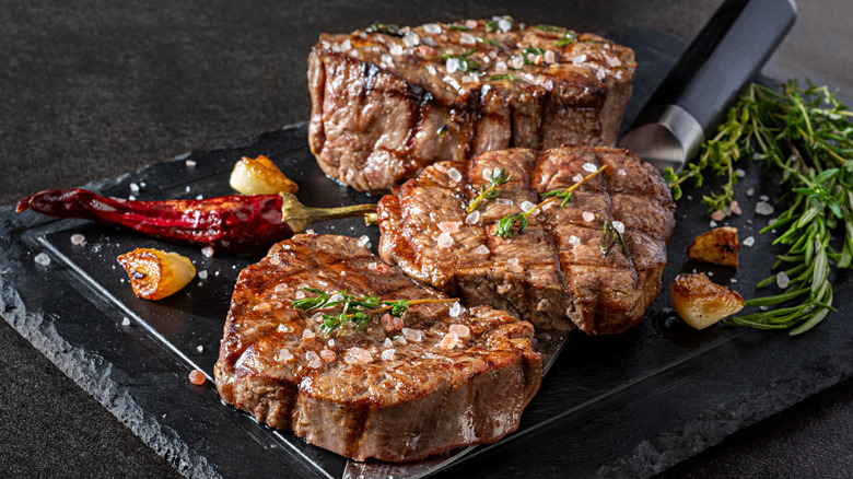 Three filets of steak on a platter, topped with salt and herbs, surrounded by a caramelized pepper and garlic, and fresh rosemary.