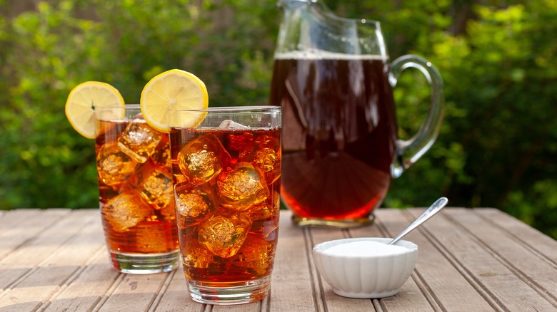 A close-up of two glasses of sweet tea on ice with lemon wheels with a bowl of sugar next to them, and a pitcher of sweet tea in the background.